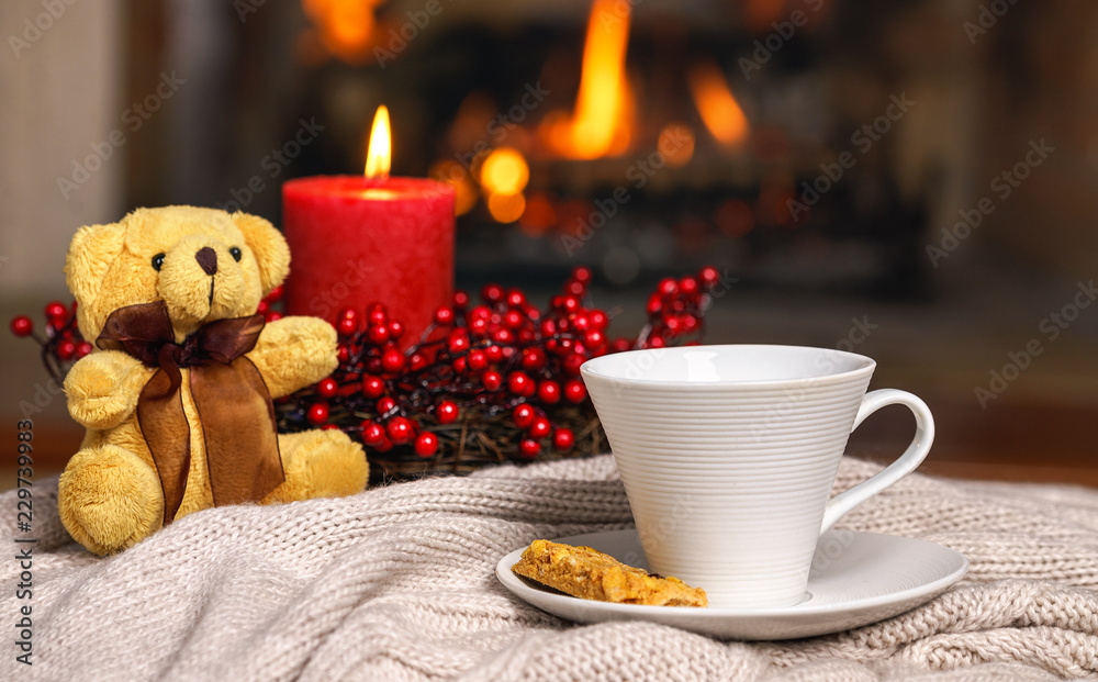 Cup of hot drink teddy bear candle in red Christmas decoration on cozy knitted plaid in front of warm fireplace. Cozy relaxed magical atmosphere in home interior. Holiday Christmas New Year concept. Cup of hot drink teddy bear candle in red Christmas decoration on cozy knitted plaid in front of warm fireplace. Cozy relaxed magical atmosphere in home interior. Holiday Christmas New Year concept.