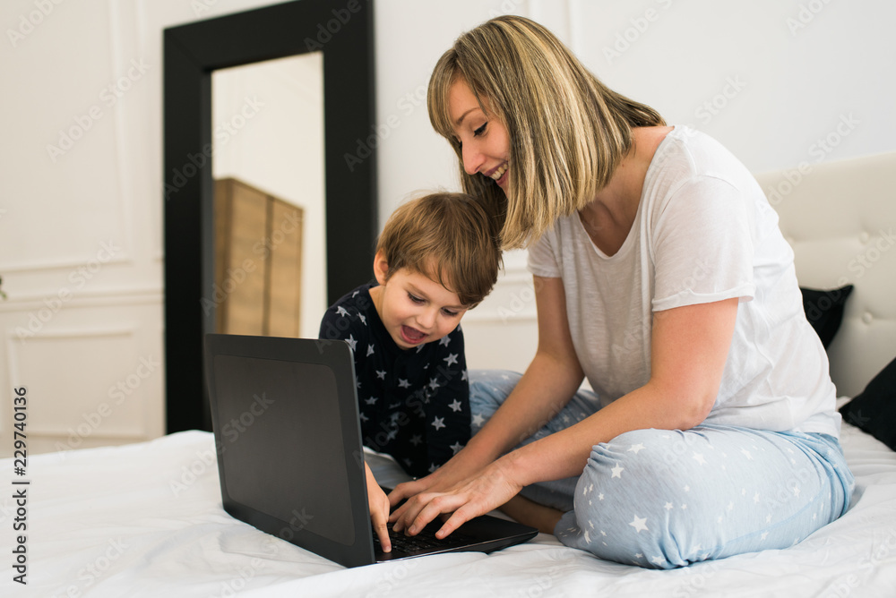 Happy mother and cute kid using laptop, sitting on the bed in pajamas and smiling. Happy mother and cute kid using laptop, sitting on the bed in pajamas and smiling.