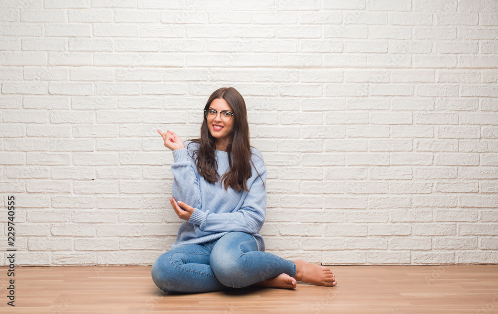Young brunette woman sitting on the floor over white brick wall very happy pointing with hand and finger to the side Young brunette woman sitting on the floor over white brick wall very happy pointing with hand and finger to the side