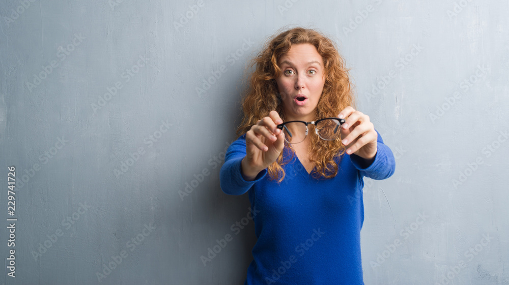 Young redhead woman over grey grunge wall holding optical glasses scared in shock with a surprise face, afraid and excited with fear expression Young redhead woman over grey grunge wall holding optical glasses scared in shock with a surprise face, afraid and excited with fear expression