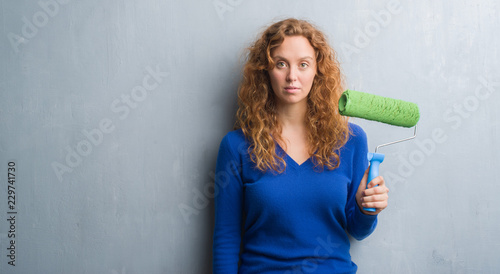 Young redhead woman over grey grunge wall holding painting roller with a confident expression on smart face thinking serious