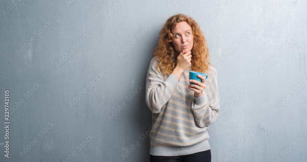 Young redhead woman over grey grunge wall drinking a cup of coffee serious face thinking about question, very confused idea Young redhead woman over grey grunge wall drinking a cup of coffee serious face thinking about question, very confused idea