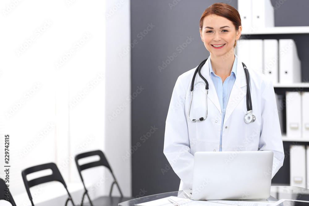 Happy doctor woman at work. Portrait of female physician using laptop computer while standing near reception desk at clinic or emergency hospital. Medicine concept Happy doctor woman at work. Portrait of female physician using laptop computer while standing near reception desk at clinic or emergency hospital. Medicine concept