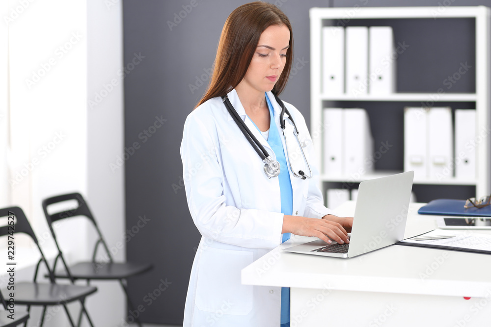 Doctor woman at work. Portrait of female physician using laptop computer while standing near reception desk at clinic or emergency hospital. Medicine and healthcare concept Doctor woman at work. Portrait of female physician using laptop computer while standing near reception desk at clinic or emergency hospital. Medicine and healthcare concept