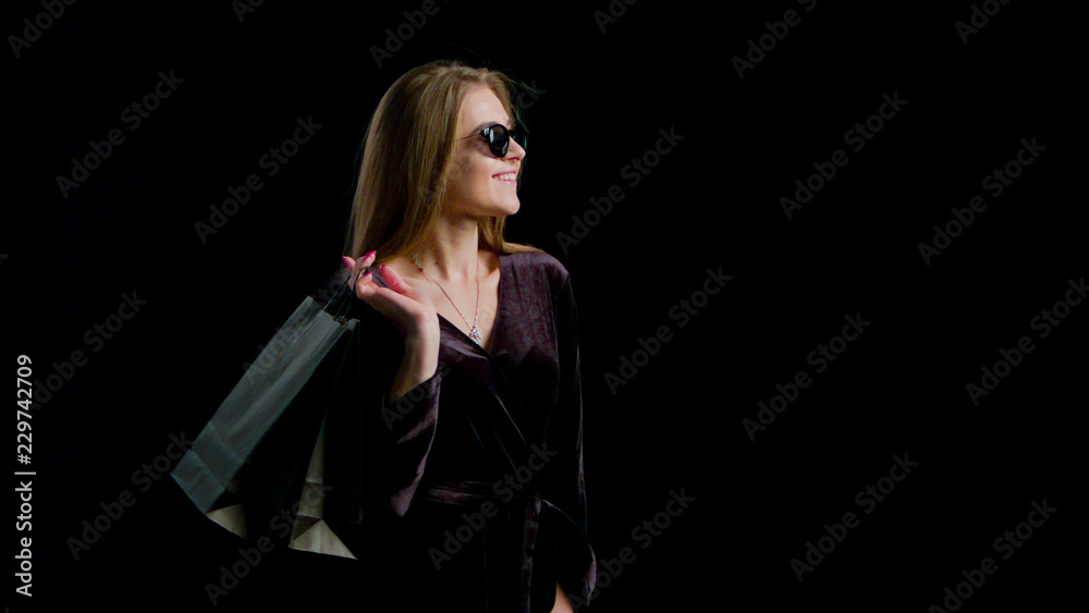 Woman in cool sunglasses and black dress, holding black shopping bag isolated on dark background in black friday holiday or christmas Woman in cool sunglasses and black dress, holding black shopping bag isolated on dark background in black friday holiday or christmas