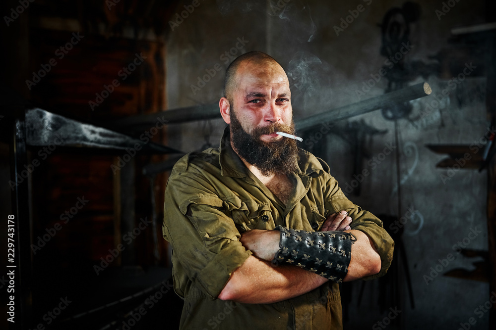 Brutal bearded blacksmith worker smokes after hard work. The man is tired, but smiling, his face is covered in mud and sweat Brutal bearded blacksmith worker smokes after hard work. The man is tired, but smiling, his face is covered in mud and sweat