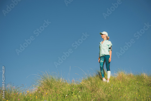 Fitness runner woman listening to music on the nature. Portrait of beautiful girl wearing earphones earbuds and running cap. against the background of a green field and blue sky © yanik88