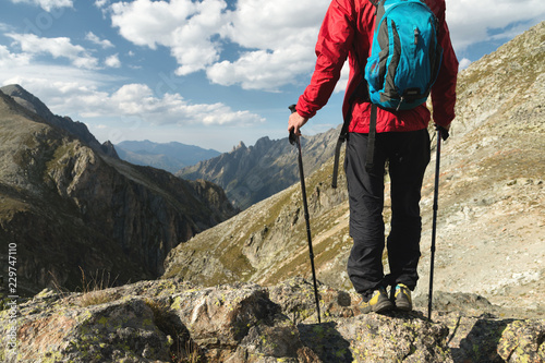 The body of man with a backpack and trekking poles stands on top of a rock against the background rocky valley high in the mountains. The concept of tourism and easy trekking in the mountains outdoor