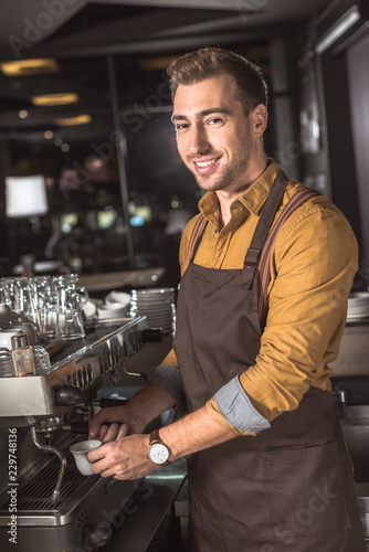 handsome young barista preparing coffee with coffee machine in cafe and looking at camera
