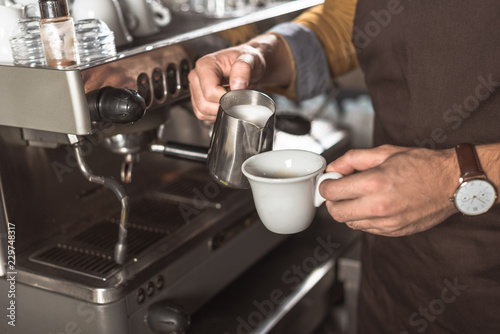 cropped shot of barista in apron pouring milk into coffee while preparing it in cafe