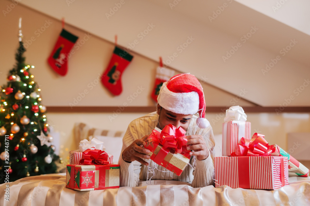 Fototapeta premium Mixed race man with santa's hat on head lying on bed and trying to open Christmas gift. Around him presents, in background Christmas decorations.