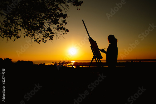 silhouette of a woman painting a picture with paints on canvas on an easel, girl with paint brush and palette engaged in art on the nature in a field at sunset © fantom_rd