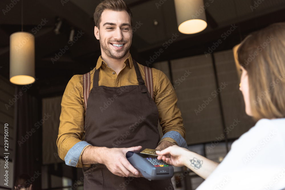 smiling young waiter holding payment terminal while customer doing contactless purchase with credit card in cafe