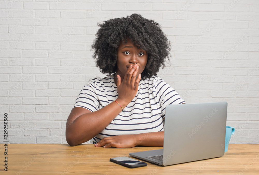 Young african american woman sitting on the table using computer laptop cover mouth with hand shocked with shame for mistake, expression of fear, scared in silence, secret concept