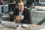 handsome young freelancer with cup of coffee and laptop sitting in cafe