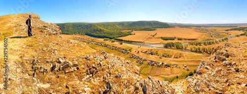 Man Tourist with a backpack is on top of the High Rock Mountain below, the Zilyim River flows against the background of the Ural Mountains. Autumn sunny day. photo