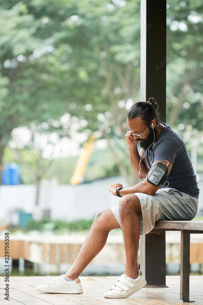 Athletic bearded Indian man in sportswear sitting on bench outdoors and  looking at his watch while taking pulse after running Stock Photo | Adobe  Stock