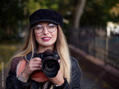 Portrait of a beautiful stylish cheerful young blonde photographer wearing glasses and a cap with makeup holding a reflex camera in her hands in autumn park © mak_alexandra