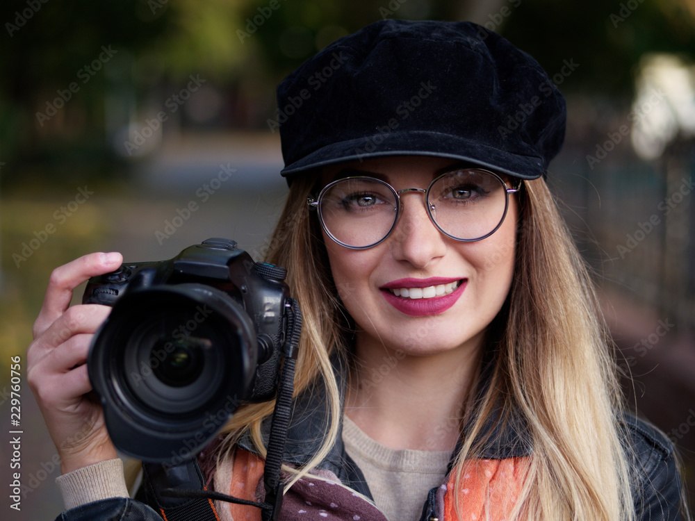 Portrait of a beautiful stylish cheerful young blonde photographer wearing glasses and a cap with makeup holding a reflex camera in her hands in autumn park