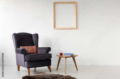 Empty wooden frame on white wall of elegant living room with comfortable armchair with patterned pillow, fury blanket and small table with book and tea © Photographee.eu