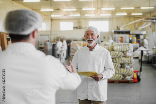 Picture of mature cheerful older man in sterile clothes standing in food factory and being happy about seeing his colleague. Team work and good job concept.