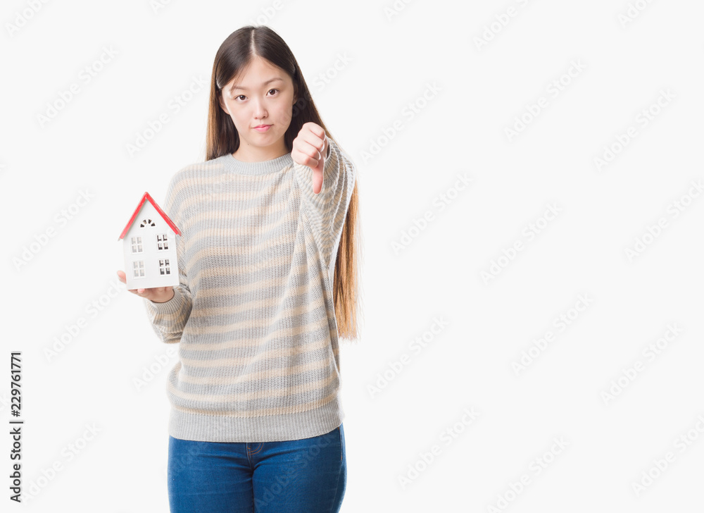 Young Chinese real state agent woman over isolated background holding house with angry face, negative sign showing dislike with thumbs down, rejection concept