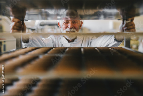 Picture of professional mature male baker man in white work uniform looking at the camera. Standing in front of the shelves full with fresh baked cookies.