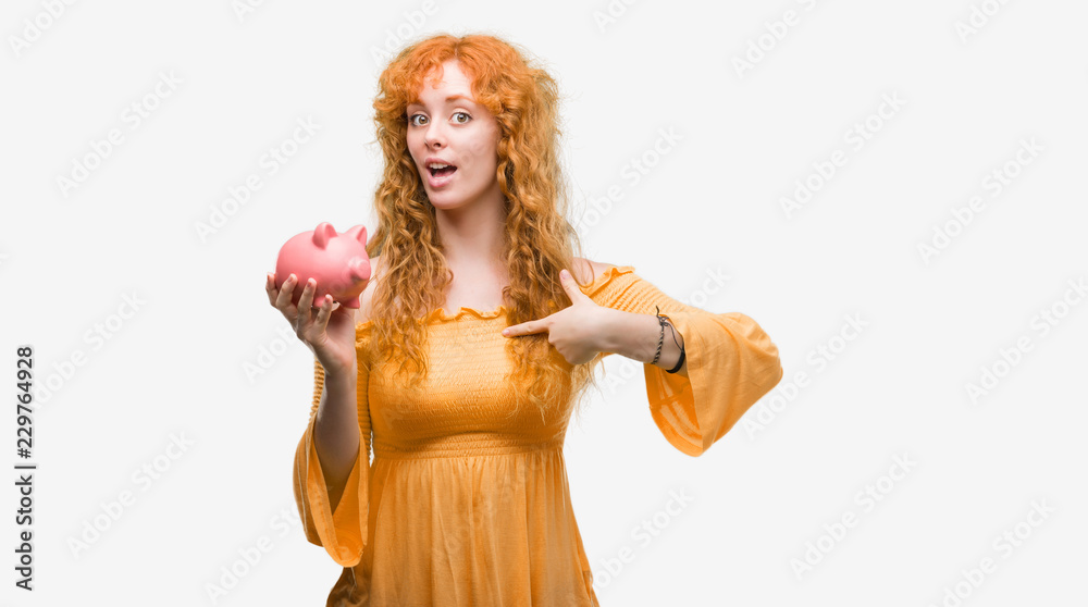 Young redhead woman holding piggy bank with surprise face pointing finger to himself