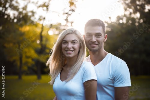Cute loving couple standing on grass in nature green park with beautiful sundown light. Looking camera.