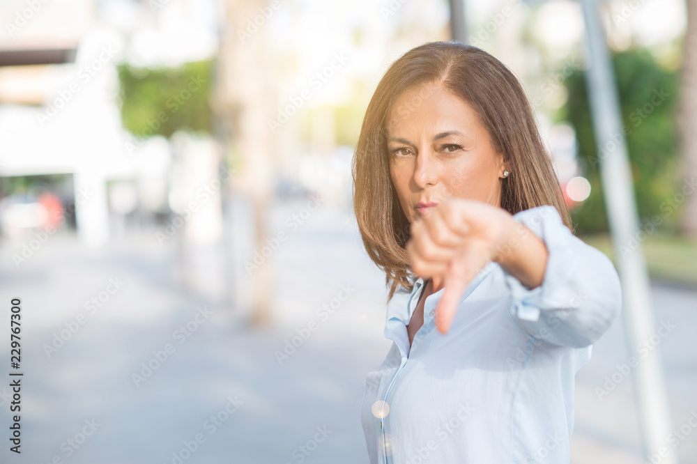 Beautiful middle age hispanic woman at the city street on a sunny day with angry face, negative sign showing dislike with thumbs down, rejection concept