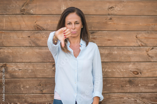 Beautiful middle age hispanic woman at over wood wall on a sunny day with angry face, negative sign showing dislike with thumbs down, rejection concept