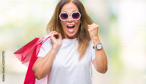 Middle age hispanic woman holding shopping bags on sales over isolated background screaming proud and celebrating victory and success very excited, cheering emotion