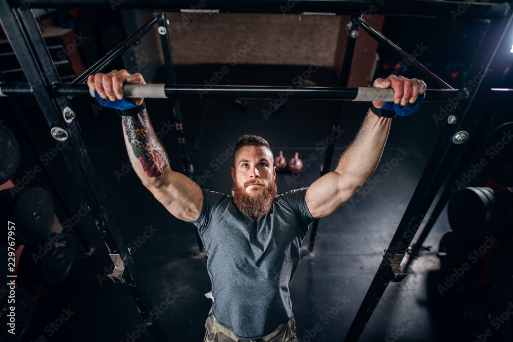 Muscular caucasian bearded man doing pull-ups and training his biceps and back in crossfit gym.