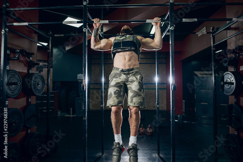 Muscular caucasian bearded man doing pull-ups in military style weighted vest in crossfit gym. Weight plates, kettlebells, barbell and crossfit tires in background.