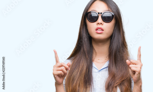 Young asian woman wearing sunglasses over isolated background amazed and surprised looking up and pointing with fingers and raised arms. © Krakenimages.com