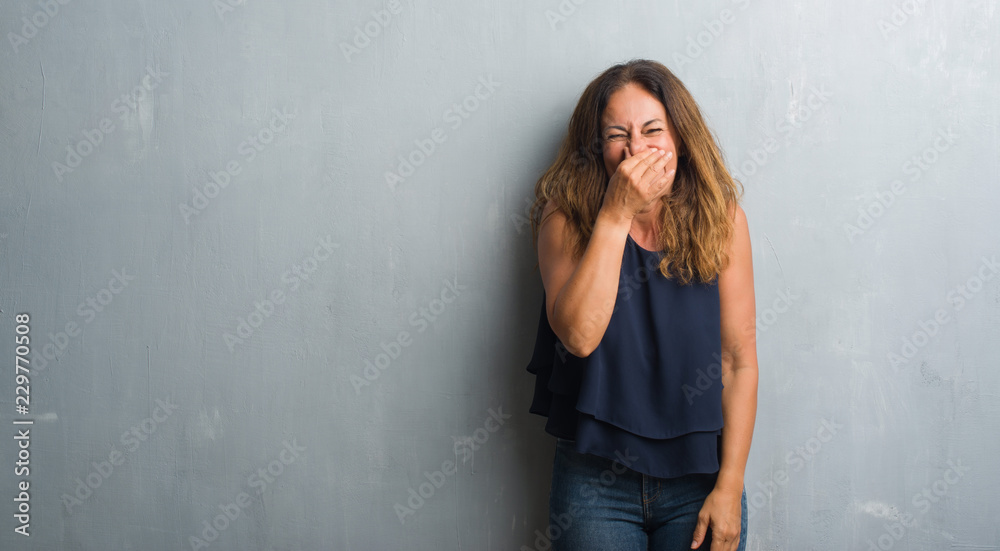Middle age hispanic woman standing over grey grunge wall smelling something stinky and disgusting, intolerable smell, holding breath with fingers on nose. Bad smells concept. Middle age hispanic woman standing over grey grunge wall smelling something stinky and disgusting, intolerable smell, holding breath with fingers on nose. Bad smells concept.