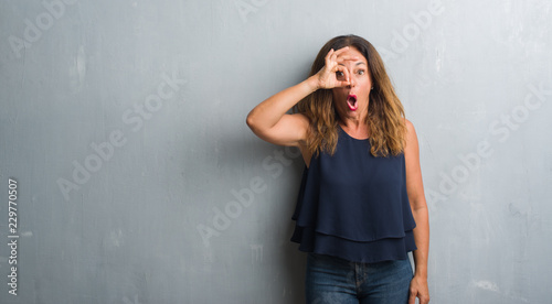 Middle age hispanic woman standing over grey grunge wall doing ok gesture shocked with surprised face, eye looking through fingers. Unbelieving expression.