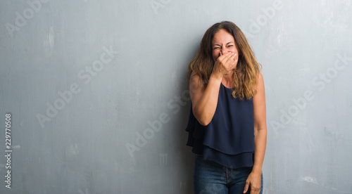 Middle age hispanic woman standing over grey grunge wall smelling something stinky and disgusting, intolerable smell, holding breath with fingers on nose. Bad smells concept.