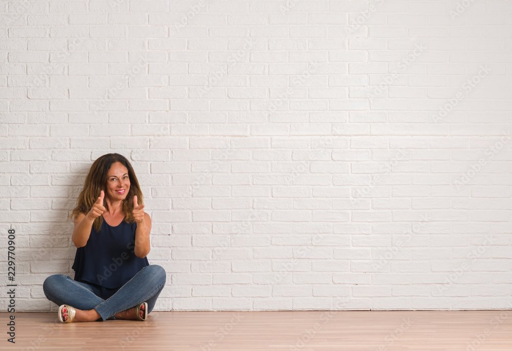 Middle age hispanic woman sitting on the floor over white brick wall pointing fingers to camera with happy and funny face. Good energy and vibes. Middle age hispanic woman sitting on the floor over white brick wall pointing fingers to camera with happy and funny face. Good energy and vibes.