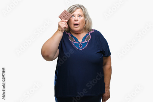 Senior plus size caucasian woman eating chocolate bar over isolated background scared in shock with a surprise face, afraid and excited with fear expression