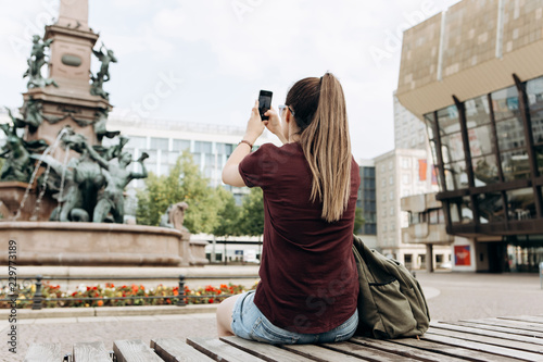 The girl on the street takes pictures of the beautiful fountain and buildings in Leipzig in Germany. She makes a street mobile photo to share it on social networks with friends.