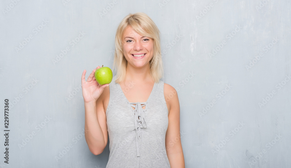 Adult caucasian woman over grunge grey wall eating green apple with a happy face standing and smiling with a confident smile showing teeth Adult caucasian woman over grunge grey wall eating green apple with a happy face standing and smiling with a confident smile showing teeth