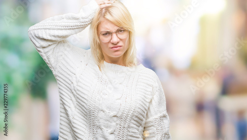 Young beautiful blonde woman wearing winter sweater and glasses over isolated background confuse and wonder about question. Uncertain with doubt, thinking with hand on head. Pensive concept.