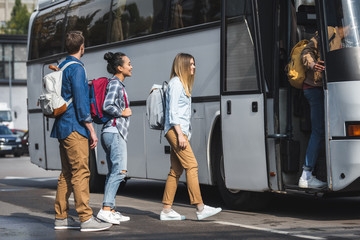 young smiling tourists with rucksacks walking into travel bus at city street