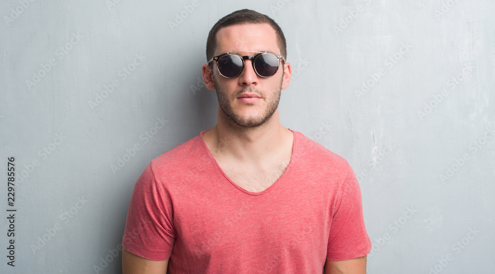 Young caucasian man over grey grunge wall wearing sunglasses with a confident expression on smart face thinking serious Young caucasian man over grey grunge wall wearing sunglasses with a confident expression on smart face thinking serious