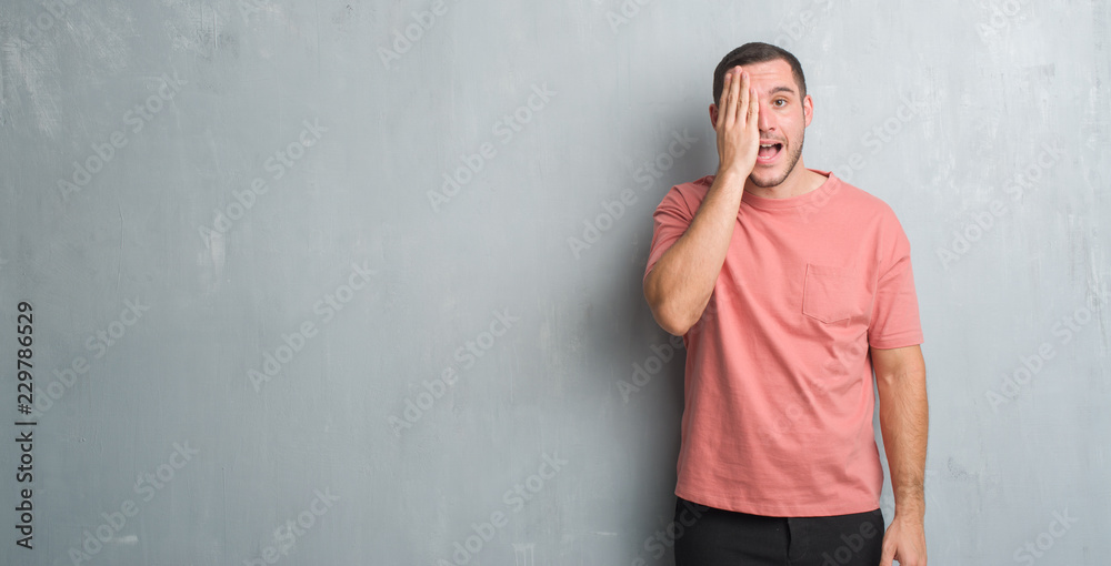 Young caucasian man over grey grunge wall covering one eye with hand with confident smile on face and surprise emotion. Young caucasian man over grey grunge wall covering one eye with hand with confident smile on face and surprise emotion.
