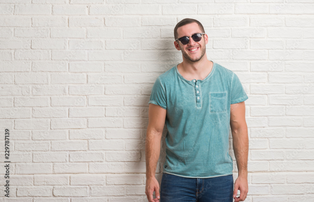 Young caucasian man standing over white brick wall wearing sunglasses with a happy face standing and smiling with a confident smile showing teeth Young caucasian man standing over white brick wall wearing sunglasses with a happy face standing and smiling with a confident smile showing teeth