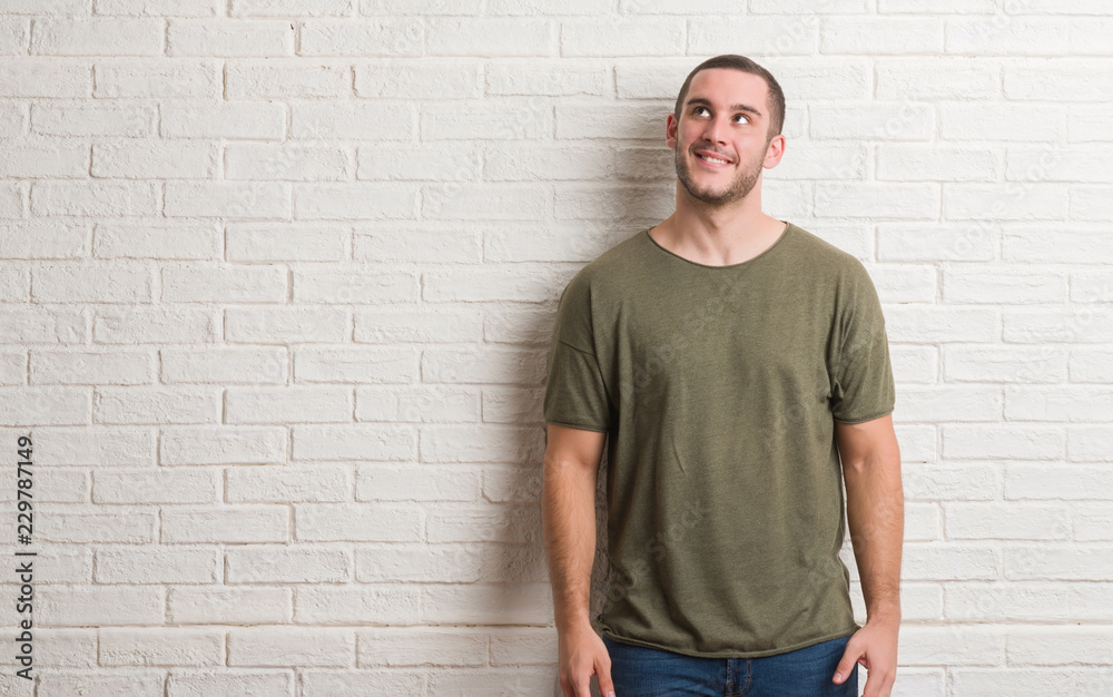 Young caucasian man standing over white brick wall smiling looking side and staring away thinking. Young caucasian man standing over white brick wall smiling looking side and staring away thinking.
