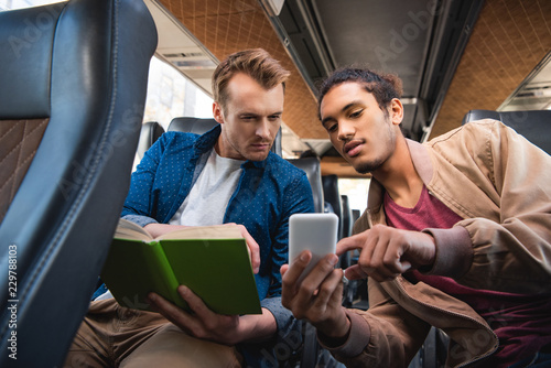 young mixed race man showing smartphone to his male friend with book during trip on travel bus