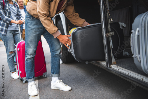 cropped image of man putting wheeled bag into travel bus while his friends standing behind at street © LIGHTFIELD STUDIOS